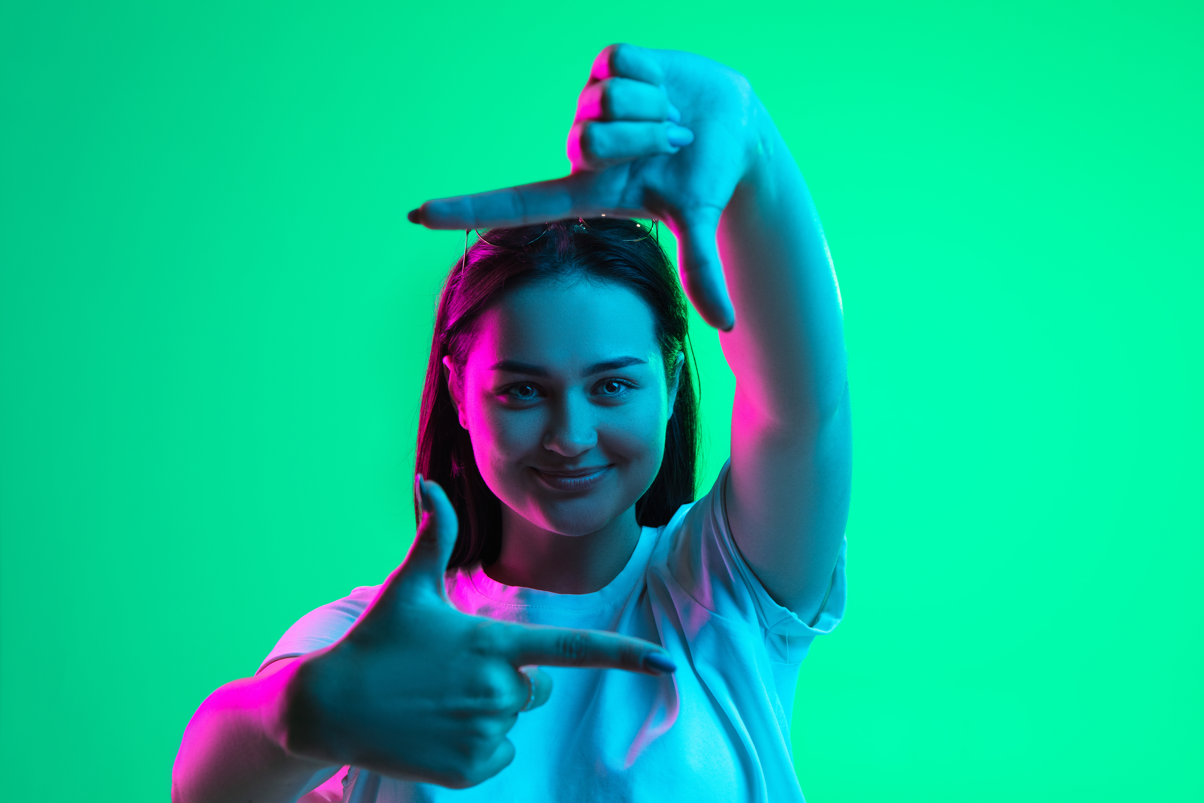 Retrato en primer plano de una joven caucásica bastante sonriente que muestra el gesto del marco aislado sobre fondo verde en luz de neón. - Foto de stock de Anuncio libre de derechos